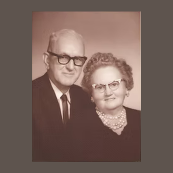 Elderly couple in vintage attire poses together, both wearing glasses. The man in a suit, the woman in a pearl necklace, conveying timeless elegance.
