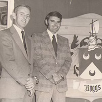 Two men in suits stand smiling in front of a stylized coat of arms featuring a ship and crescent moons, with the name "Boggs" below.