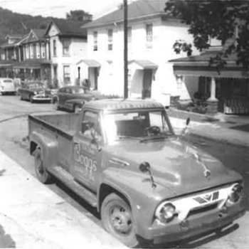 Classic pickup truck with "Boops" on the door, parked on a suburban street lined with vintage cars and houses. Black and white, nostalgic feel.