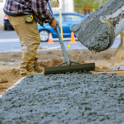 Concrete, Masonry & CompactionA worker in tan pants and plaid shirt levels wet concrete with a tool on a construction site. A blue car and orange cones are visible in the background.