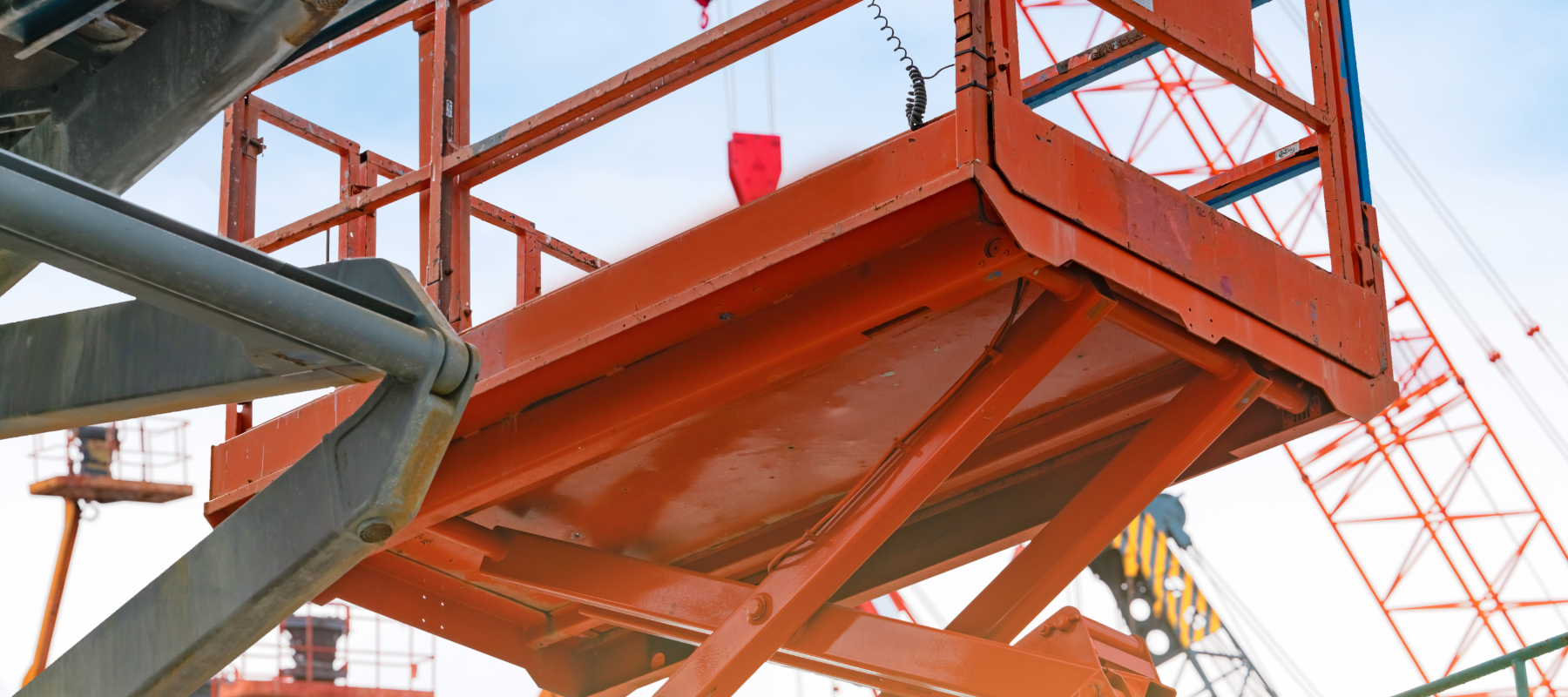 Orange construction lift with metallic framework against a clear sky, surrounded by cranes. The scene conveys a sense of industrial activity and progress.